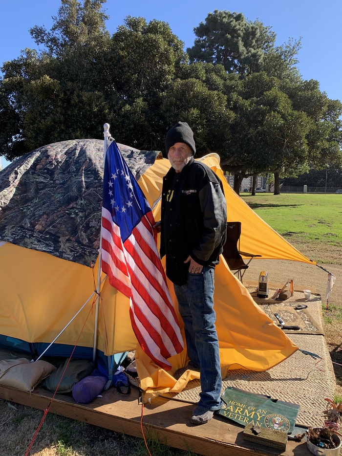 White, male, homeless veteran next to his tent and the American flag
